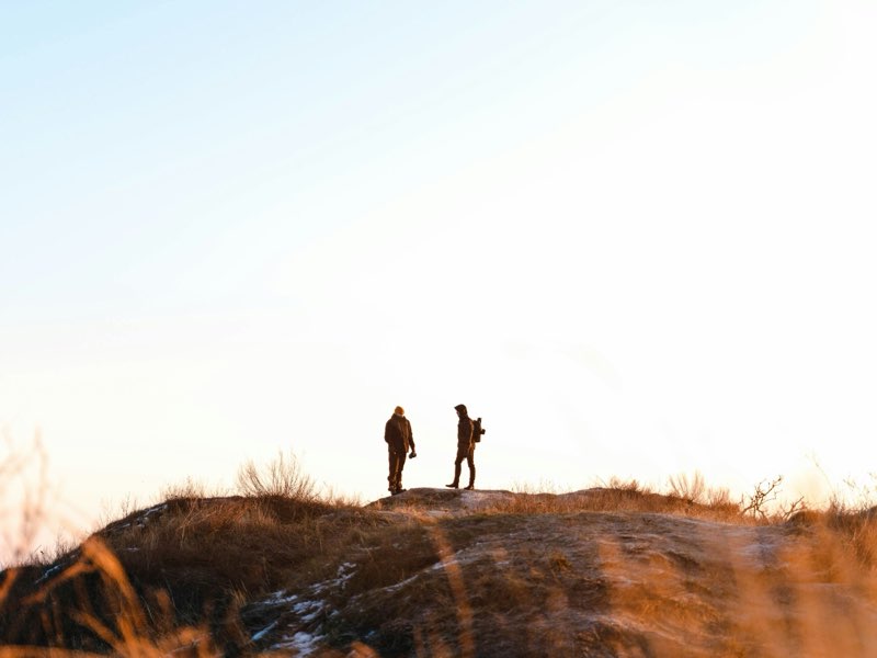 Two people, one carrying a camera, on a grassy ridge at golden hour.
